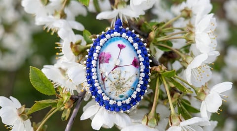 Blue and white beaded oval pendant with pressed flower design hanging among white babys breath flowers and green foliage