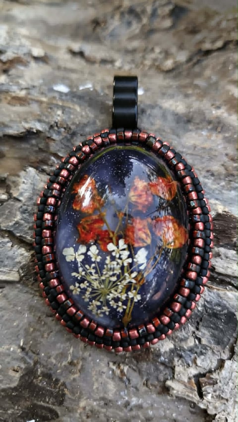 Handmade resin pendant with pressed flowers and dried botanicals embedded in dark blue resin, surrounded by copper beads, displayed on textured stone surface
