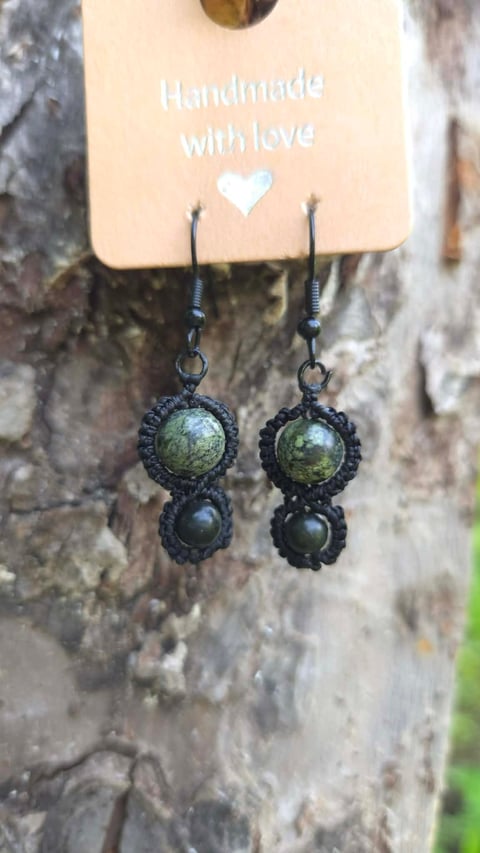 Pair of handmade dangling earrings with black ornate settings and green gemstone tops, displayed on a cream-colored tag reading Handmade with love against a weathered stone wall