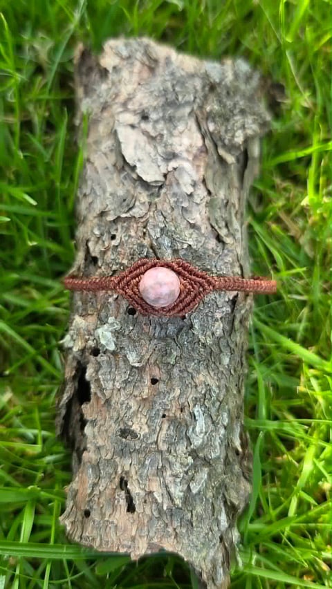 Close-up of a gray tree branch with a copper-brown wire wrapped around it, featuring a pale pink stone or bead in the center, surrounded by green grass