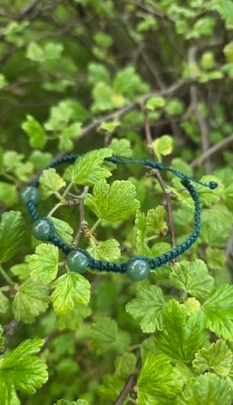 Beaded bracelet with dark blue-green cord and grey stone beads resting on green leafy plant branches
