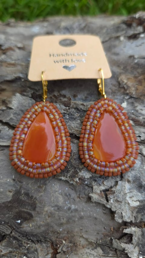 Pair of handmade teardrop earrings with orange beaded frames and amber stone centers on gold hooks, displayed on cream product tag