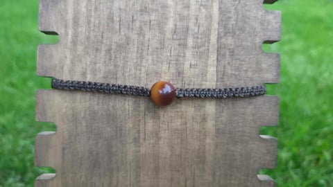 Dark braided cord bracelet with amber-colored wooden bead displayed on wooden surface with green grass background