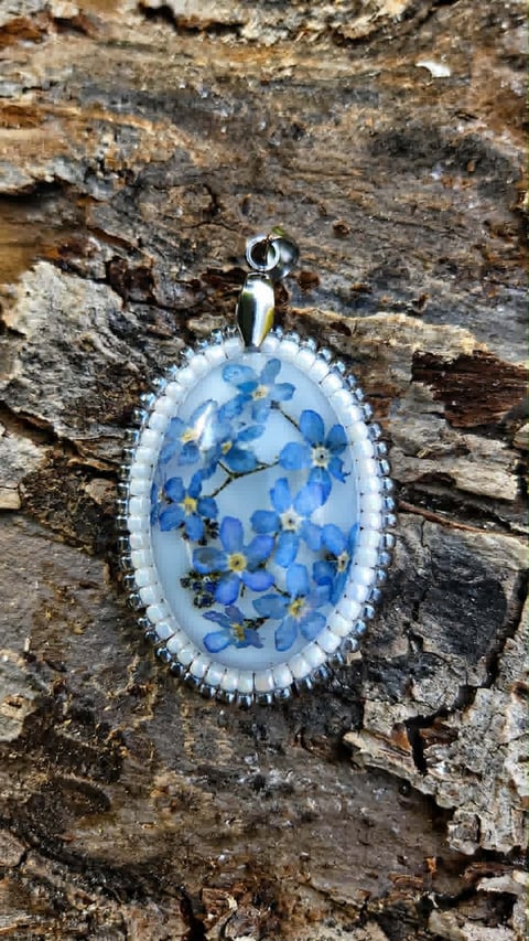 Oval resin pendant with pressed blue flowers and gold accents on beaded white border, displayed on weathered tree bark