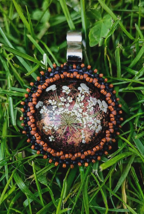 Circular resin pendant with dried flowers and orange-brown beaded border on grass background