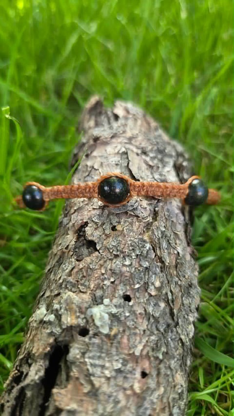 Wooden toy figure with black bead eyes and brown wire arms posed on a tree bark surrounded by green grass