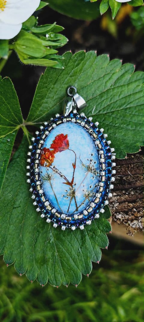 Ornate pendant with blue oval resin dome containing pressed red flower and stems, set in silver beaded frame hanging on green foliage