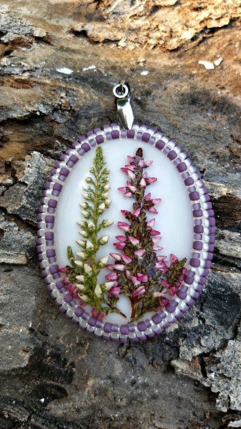 Oval resin pendant with pressed flowers featuring purple beads border, displayed on textured stone background