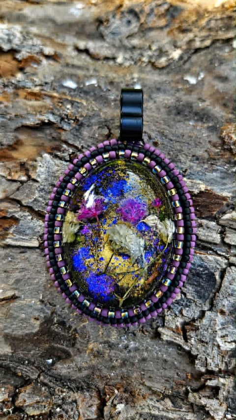 Beaded pendant with purple beads and colorful resin center featuring blue, yellow, and pink tones, displayed on textured tree bark