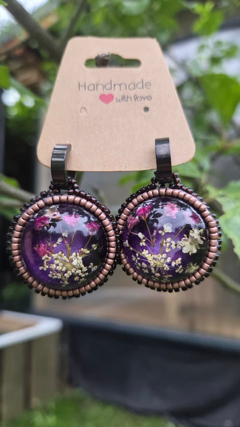 Pair of handmade drop earrings with dark purple resin domes containing dried flowers, displayed on cardstock with Handmade with love text and green foliage background