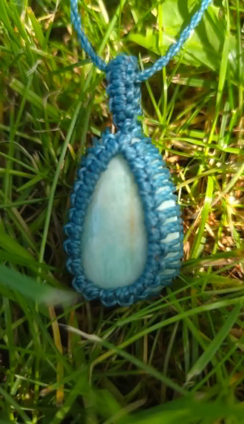 Blue teardrop-shaped pendant with pale blue stone center on blue cord hanging in green grass